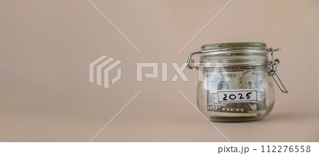 Saving Money In Glass Jar filled with Dollars banknotes. 2025 year transcription in front of jar. Managing personal finances extra income for future insecurity 112276558
