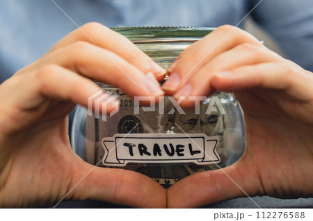 Unrecognizable woman showing heart sign Saving Money In Glass Jar filled with Dollars banknotes. TRAVEL transcription in front of jar. Managing personal finances extra income for future insecurity 112276588