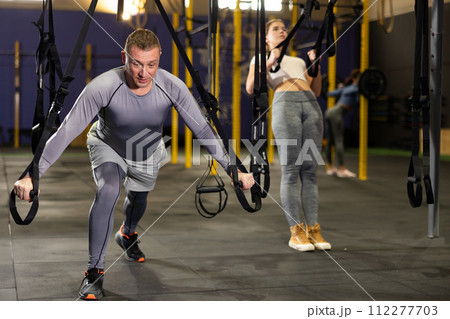 Adult male athlete with group working out on gymnastics rings at crossfit gym Adult male athlete with group working out on gymnastics rings at crossfit gym 112277703