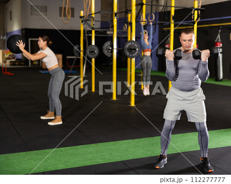 Dedicated man doing exercises with dumbbells near other people in gym Dedicated man doing exercises with dumbbells near other people in gym 112277777