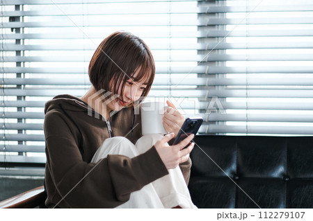 部屋着で、朝ソファーでスマホを見てリラックスしてる女性 部屋着で、朝ソファーでスマホを見てリラックスしてる女性 112279107