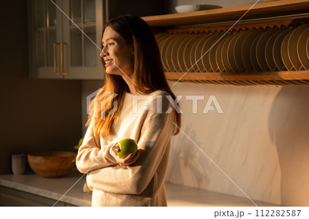 A young woman with an apple in her hands in a sunny modern kitchen at sunrise A young woman with an apple in her hands in a sunny modern kitchen at sunrise 112282587