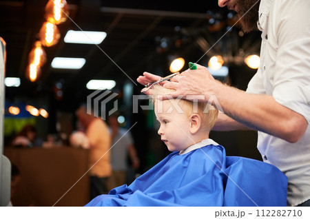 Man barber cutting little boy's hair using comb and scissors. Child getting haircut from adult male, likely barber. Professional hairdresser and cute client at modern barbershop. Man barber cutting little boy's hair using comb and scissors. Child getting haircut from adult male, likely barber. Professional hairdresser and cute client at modern barbershop. 112282710