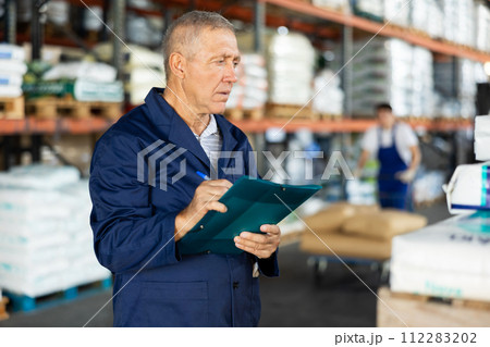 In warehouse of store, mature man checks availability of goods and receipt documents for products In warehouse of store, mature man checks availability of goods and receipt documents for products 112283202
