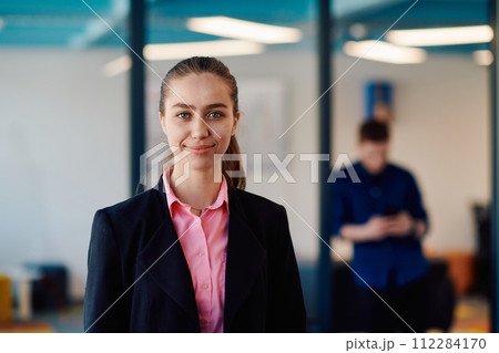 Portrait of young smiling business woman in creative open space coworking startup office. Successful businesswoman standing in office with copyspace. Coworkers working in background Portrait of young smiling business woman in creative open space coworking startup office. Successful businesswoman standing in office with copyspace. Coworkers working in background 112284170