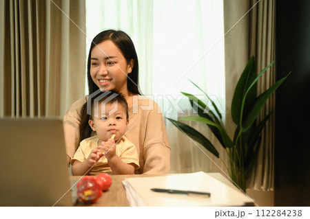 Shot of a young mother having video call with her clients over laptop and carrying her baby on laps Shot of a young mother having video call with her clients over laptop and carrying her baby on laps 112284238