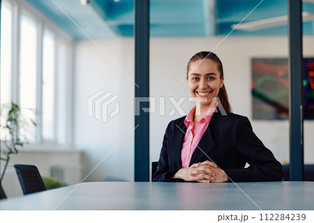Successful young female leader in a suit with a pink shirt sitting in a modern glass office with a determined smile. 112284239