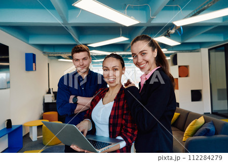 A young team of business professionals poses for a startup office portrait, exuding confidence and ambition, with one woman holding a laptop, symbolizing innovation and collaboration in 112284279
