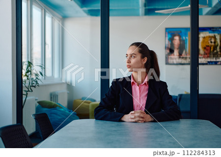Successful young female leader in a suit with a pink shirt sitting in a modern glass office with a determined smile. 112284313