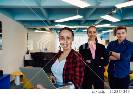 A young team of business professionals poses for a startup office portrait, exuding confidence and ambition, with one woman holding a laptop, symbolizing innovation and collaboration in A young team of business professionals poses for a startup office portrait, exuding confidence and ambition, with one woman holding a laptop, symbolizing innovation and collaboration in 112284349