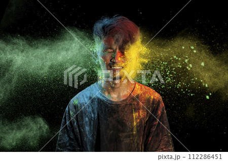 Asian man splashed with colorful holi powder and having fun isolated on black background. 112286451