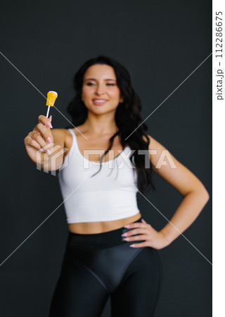 Young female sports fitness trainer holding lollypop in outstretched hand on black background 112286675