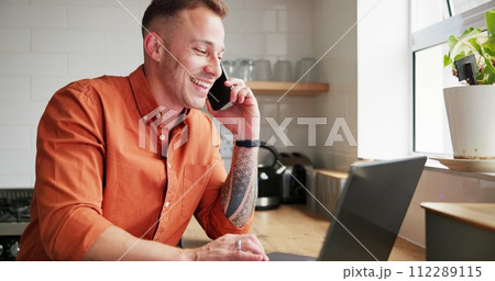 Happy man, laptop and phone call in remote work for conversation or business in kitchen at home. Male person or freelancer smile talking on mobile smartphone with computer for discussion at house 112289115