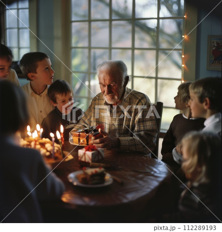 AI-generated content. An elderly man sits at a table, surrounded by smiling grandchildren. He holds a gift in his hands, while a birthday cake with lit candles is placed nearby 112289193