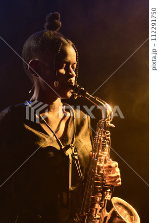 Vertical portrait of Black woman playing saxophone in dark nightclub with golden contouring light 112291750