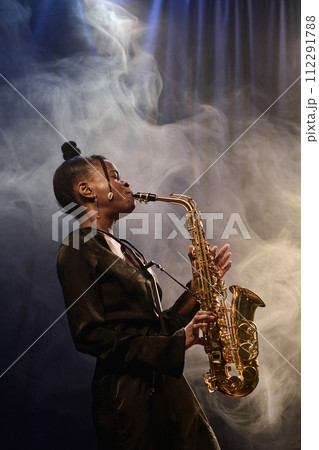 Vertical portrait of talented Black woman playing saxophone on stage during performance in jazz music club with smoke effect 112291788
