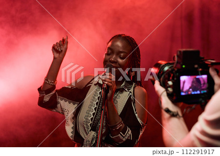 Waist up portrait of Black young woman performing in music video with unrecognizable cameraman filming 112291917