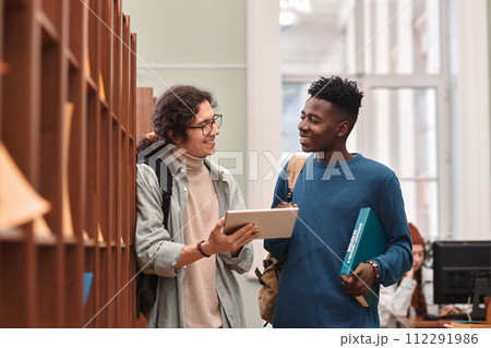 Waist up portrait of two smiling young men standing by shelves in college library and using digital tablet copy space Waist up portrait of two smiling young men standing by shelves in college library and using digital tablet copy space 112291986