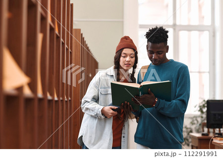 Waist up portrait of two students young man and woman reading book together standing by bookshelf in college library copy space 112291991
