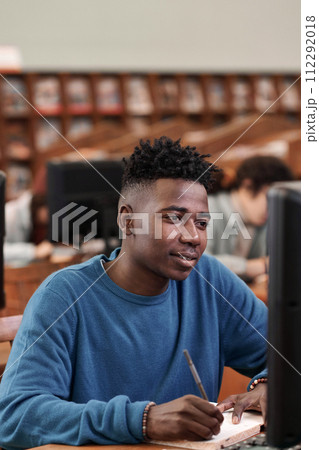 Vertical portrait of smiling young Black man looking at computer screen while doing independent study in college library 112292018