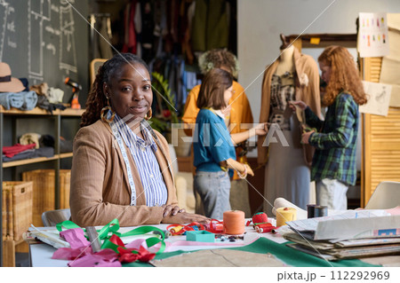 Portrait of African American adult woman sitting at desk in atelier workshop and looking at camera with girls in background copy space 112292969