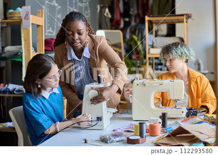 Portrait of Black young woman teaching girl with disability using sewing machines in vocational training class copy space 112293580