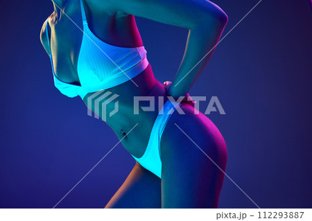 Close-up of woman in underwear, showcasing belly with smooth and toned skin in neon light against blue studio background 112293887
