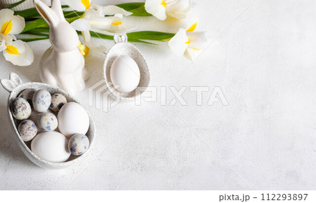 Easter white holiday card. Natural colors eggs in figured bowls, ceramic bunny, flowers. Copy space. 112293897