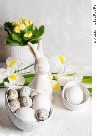 White holiday decorations on Easter table. Eggs in bowls with bunny ears, ceramic bunny, flowers. White holiday decorations on Easter table. Eggs in bowls with bunny ears, ceramic bunny, flowers. 112293898