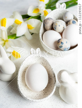 Easter natural colors eggs in bowls with bunny ears, bunnies, white Iris flowers on white close up. Easter natural colors eggs in bowls with bunny ears, bunnies, white Iris flowers on white close up. 112293917
