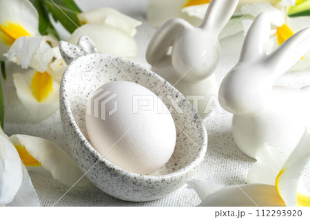 One Easter egg in marble bowl with bunny ears, small bunnies, white Iris flowers on white close up. 112293920