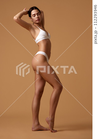 Side view portrait of young woman with slim figure posing in white underwear raising arms against sandy color studio background. Side view portrait of young woman with slim figure posing in white underwear raising arms against sandy color studio background. 112293946