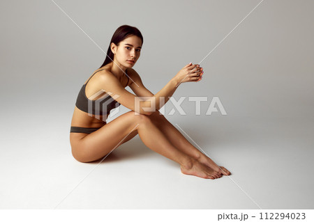 Young, tender woman with slim body posing in lingerie sitting on floor and looking at camera against grey studio background. 112294023