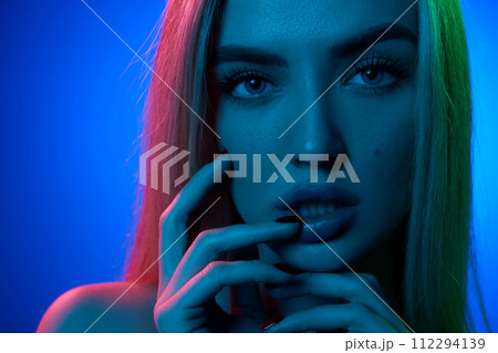 Portrait of attractive, sensual, beautiful young woman with blonde hair, plump lips and perfect skin against blue studio background in neon light 112294139