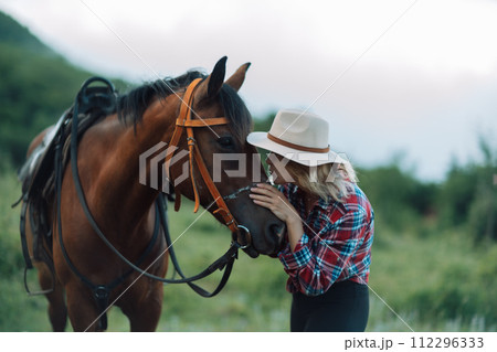 Happy blonde with horse in forest. Woman and a horse walking through the field during the day. Dressed in a plaid shirt and black leggings. 112296333