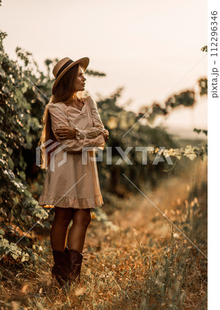 Woman with straw hat stands in front of vineyard. She is wearing a light dress and posing for a photo. Travel concept to different countries 112296346