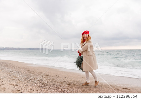 Redhead woman Christmas tree sea. Christmas portrait of a happy redhead woman walking along the beach and holding a Christmas tree in her hands. She is dressed in a light coat and a red beret. Redhead woman Christmas tree sea. Christmas portrait of a happy redhead woman walking along the beach and holding a Christmas tree in her hands. She is dressed in a light coat and a red beret. 112296354