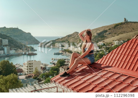 Woman sits on rooftop, enjoys town view and sea mountains. Peaceful rooftop relaxation. Below her, there is a town with several boats visible in the water. Rooftop vantage point. Woman sits on rooftop, enjoys town view and sea mountains. Peaceful rooftop relaxation. Below her, there is a town with several boats visible in the water. Rooftop vantage point. 112296398