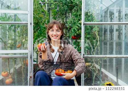 Young smiling woman holding harvest of ripe red beef tomatoes, just picked in green house. Urban farming lifestyle. Cottage core. Growing organic vegetables in garden. Concept of food self-sufficiency 112298331