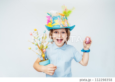 Easter eggs hunt tradition. Emotionally shouting boy kid in Easter hat holds easter tree in pot and shaking chocolate egg in other hand on the white background. Easter decoration and traditions, 112298358