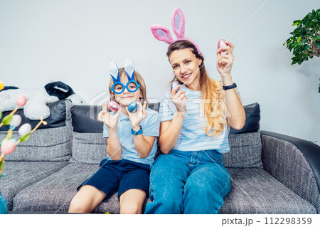 Portraits of happy smiling Caucasian mother and her son in bunny ears having fun with easter eggs at home on Easter day. Family sitting on sofa at home. Family Easter traditions. Selective focus. 112298359