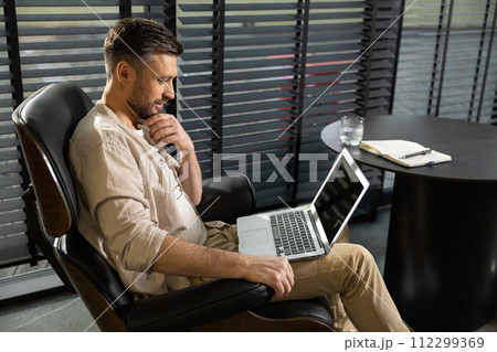 Young bearded man in casual clothes working on laptop Young bearded man in casual clothes working on laptop 112299369