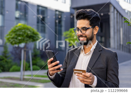 A cheerful Indian man stands outside an office complex, expertly handling a smartphone in one hand and a credit card in the other. The image exudes secure financial transactions. 112299666
