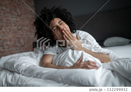 Curly-haired young man lying in bed and yawning Curly-haired young man lying in bed and yawning 112299932