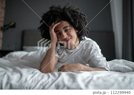 Young curly-haired man lying in bed and looking relaxed and sleepy 112299940