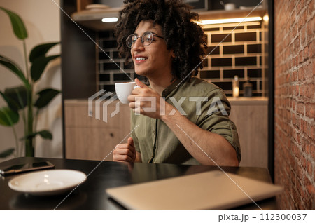 Curly-haired young man sitting at the table and having coffee Curly-haired young man sitting at the table and having coffee 112300037