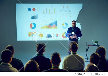 Presenter with ID badge stands in front of an audience, giving lecture with data chart backdrop in conference room. 112303325