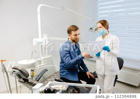 Young attractive man visiting dentist, sitting in dental chair at modern light clinic. Young woman dentist holding x ray image. 112304898