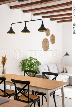 Vertical shot of a kitchen with a wooden table and chairs against the backdrop of a living room with loft lamps and a beamed ceiling. Scandinavian interior concept in a new building Vertical shot of a kitchen with a wooden table and chairs against the backdrop of a living room with loft lamps and a beamed ceiling. Scandinavian interior concept in a new building 112305353