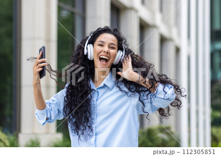 An energetic young woman with long curly hair, enjoying her favorite tunes with headphones, holding a smartphone outdoors with office buildings in the background, conveying a sense freedom and joy. An energetic young woman with long curly hair, enjoying her favorite tunes with headphones, holding a smartphone outdoors with office buildings in the background, conveying a sense freedom and joy. 112305851
